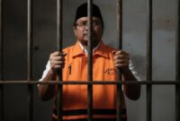 Ilustrasi Eks Menag Yaqut Cholil Qoumas mengenakan rompi oranye di dalam sel tahanan.
