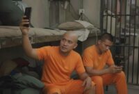 Ilustrasi narapidana menggunakan handphone di dalam kamar hunian Lapas, menggambarkan dugaan kelalaian pengawasan di lapas.