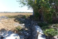 Kondisi proyek rehabilitasi jaringan irigasi Daerah Irigasi (DI) Pamukkulu di Kabupaten Takalar yang mangkrak dan belum rampung, memperlihatkan material batu serta struktur saluran yang terbengkalai di area persawahan.
