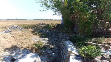 Kondisi proyek rehabilitasi jaringan irigasi Daerah Irigasi (DI) Pamukkulu di Kabupaten Takalar yang mangkrak dan belum rampung, memperlihatkan material batu serta struktur saluran yang terbengkalai di area persawahan.