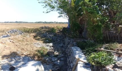 Kondisi proyek rehabilitasi jaringan irigasi Daerah Irigasi (DI) Pamukkulu di Kabupaten Takalar yang mangkrak dan belum rampung, memperlihatkan material batu serta struktur saluran yang terbengkalai di area persawahan.