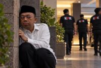 Ilustrasi dramatis Nusron Wahid yang digambarkan duduk bersembunyi di balik tembok sambil mengawasi situasi di sekitarnya. (Visual AI).